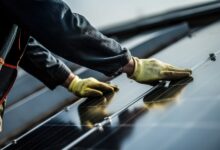 A worker wearing gloves is installing solar panels on a roof, with mounting rails and cloudy sky nearby.