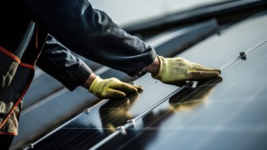 A worker wearing gloves is installing solar panels on a roof, with mounting rails and cloudy sky nearby.