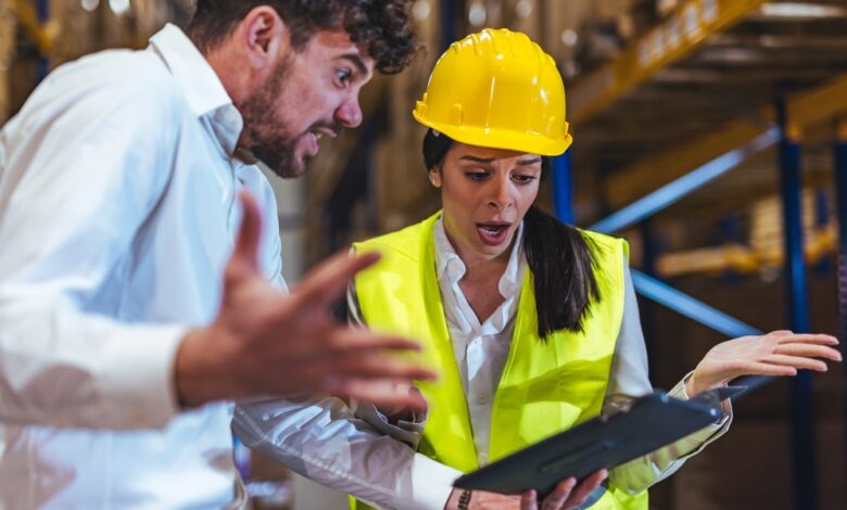 Two workers wearing hard hats and safety vests disagreeing while reviewing paperwork beside warehouse shelves.