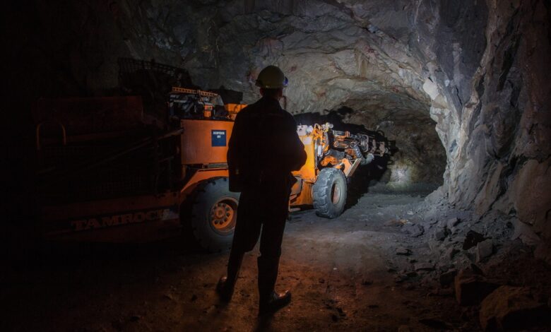 A person in a hard hat stands in a rocky underground tunnel beside an orange scoop machine and scattered stones.