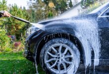 A man using a pressure washer to spray white foam on a black car in a garden with greenery and plants nearby.