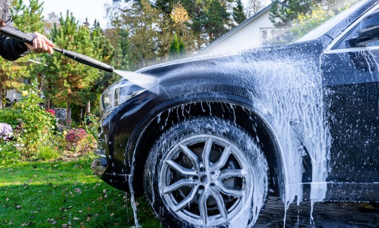 A man using a pressure washer to spray white foam on a black car in a garden with greenery and plants nearby.