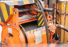 An orange industrial winch holds a coiled steel cable on a rig floor with striped guards and metal railings nearby.