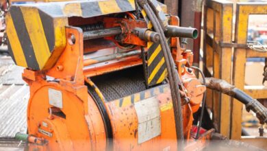 An orange industrial winch holds a coiled steel cable on a rig floor with striped guards and metal railings nearby.