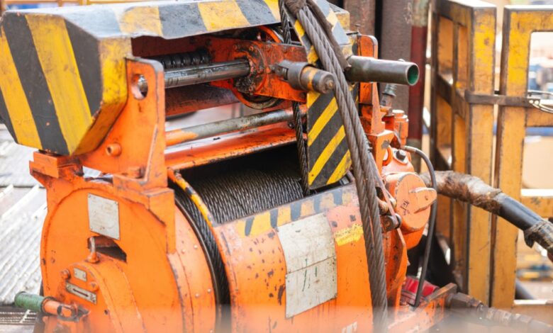 An orange industrial winch holds a coiled steel cable on a rig floor with striped guards and metal railings nearby.
