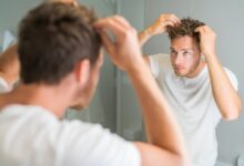 A man in a white T-shirt examining and adjusting his hair with both hands while looking into a bathroom mirror.
