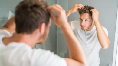 A man in a white T-shirt examining and adjusting his hair with both hands while looking into a bathroom mirror.