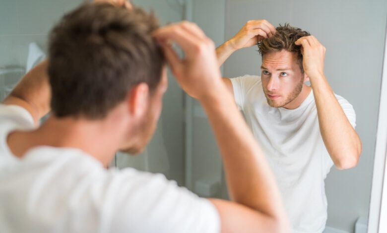 A man in a white T-shirt examining and adjusting his hair with both hands while looking into a bathroom mirror.