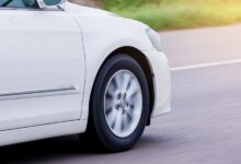 A white car speeds along a paved road as the front wheel and silver spokes blur against a sunlit street background.