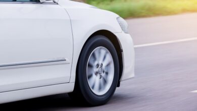 A white car speeds along a paved road as the front wheel and silver spokes blur against a sunlit street background.