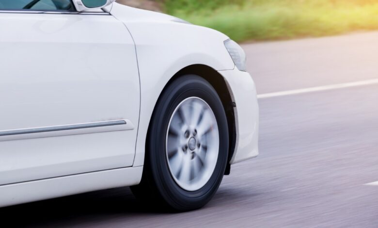 A white car speeds along a paved road as the front wheel and silver spokes blur against a sunlit street background.