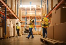 Warehouse workers in safety vests and hard hats moving boxes and checking inventory between stocked shelves.