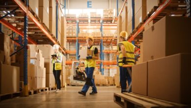 Warehouse workers in safety vests and hard hats moving boxes and checking inventory between stocked shelves.