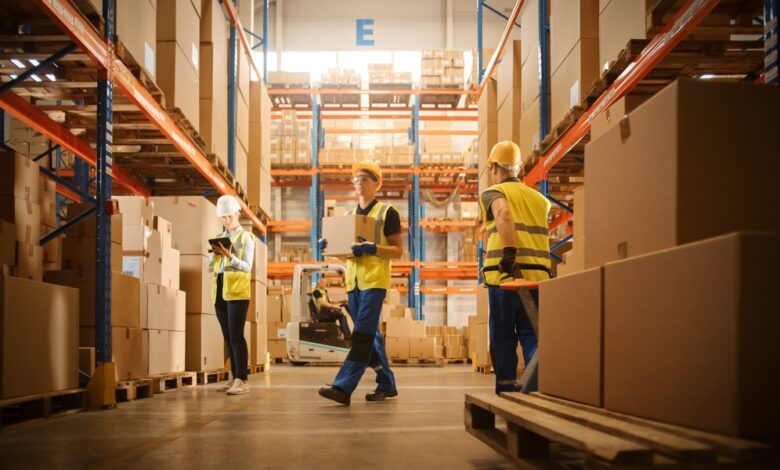 Warehouse workers in safety vests and hard hats moving boxes and checking inventory between stocked shelves.