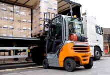 A rear view of a forklift lifting a pallet loaded with stacks of cardboard boxes wrapped in stretch film into a semi-truck.