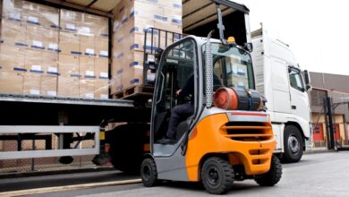 A rear view of a forklift lifting a pallet loaded with stacks of cardboard boxes wrapped in stretch film into a semi-truck.