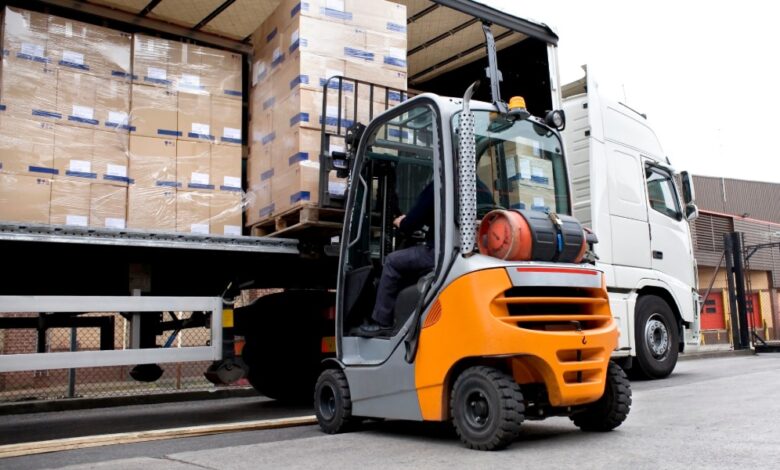 A rear view of a forklift lifting a pallet loaded with stacks of cardboard boxes wrapped in stretch film into a semi-truck.