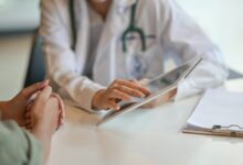 A doctor is sitting at a desk. There is another person also sitting at the desk as the doctor hold up a tablet.