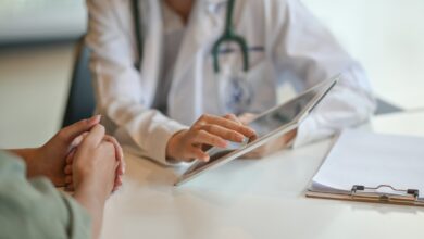 A doctor is sitting at a desk. There is another person also sitting at the desk as the doctor hold up a tablet.