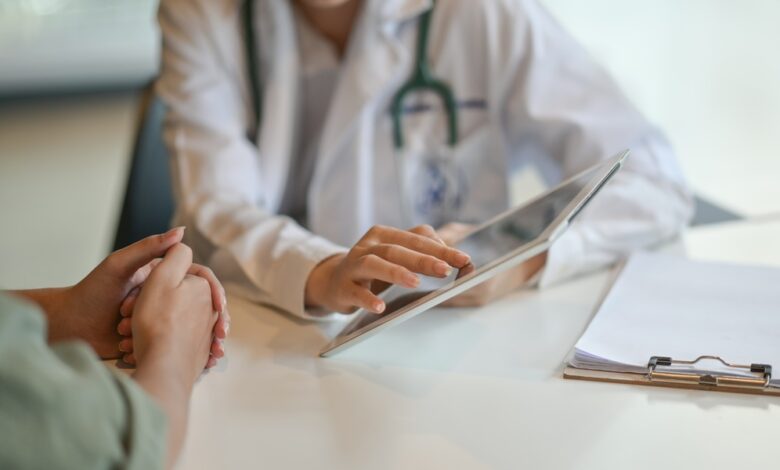 A doctor is sitting at a desk. There is another person also sitting at the desk as the doctor hold up a tablet.