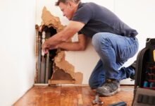 A man kneeling next to a large hole on the edge of a white wall, working on multiple pipes placed inside.