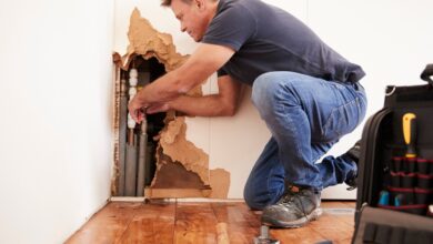 A man kneeling next to a large hole on the edge of a white wall, working on multiple pipes placed inside.
