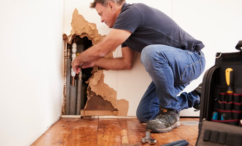 A man kneeling next to a large hole on the edge of a white wall, working on multiple pipes placed inside.