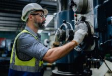 A man in a hardhat is working on a piece of industrial equipment. He’s holding a large valve and is wearing gloves.