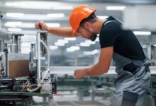 A man in an orange hard hat is leaning over a machine. He’s in an industrial environment.