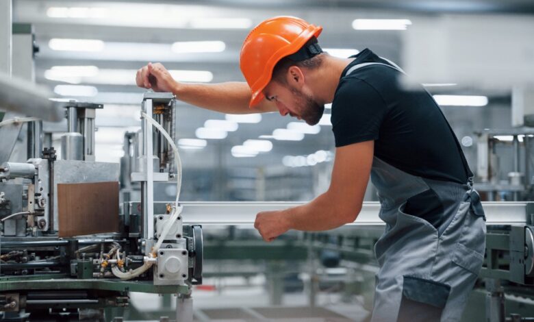 A man in an orange hard hat is leaning over a machine. He’s in an industrial environment.