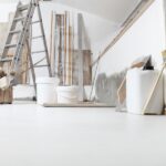A room with white floors, an unfinished painted wall, a ladder, buckets, thin wooden beams, brushes, and spatulas.