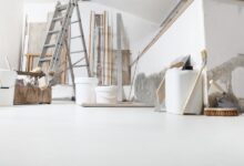 A room with white floors, an unfinished painted wall, a ladder, buckets, thin wooden beams, brushes, and spatulas.