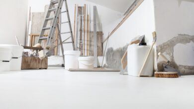A room with white floors, an unfinished painted wall, a ladder, buckets, thin wooden beams, brushes, and spatulas.