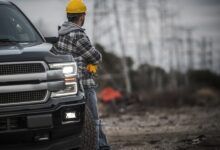 A man wearing a yellow helmet leaning onto a black pickup truck with the headlights on parked on a dirt road.