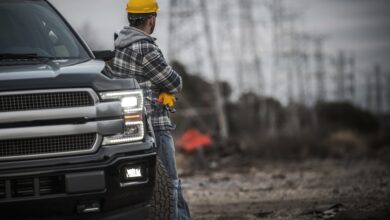 A man wearing a yellow helmet leaning onto a black pickup truck with the headlights on parked on a dirt road.