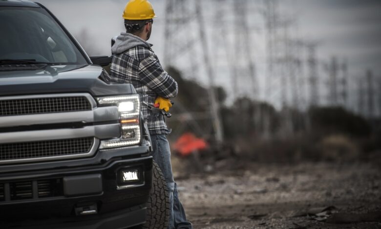 A man wearing a yellow helmet leaning onto a black pickup truck with the headlights on parked on a dirt road.