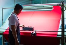 A man standing in front of a machine holding a large sheet of red textile, pushing a button on a remote control.
