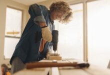 Worker wearing gloves uses a cordless drill on a piece of wood at a workbench inside a bright room under renovation.