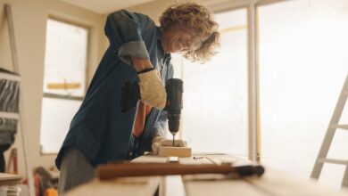 Worker wearing gloves uses a cordless drill on a piece of wood at a workbench inside a bright room under renovation.