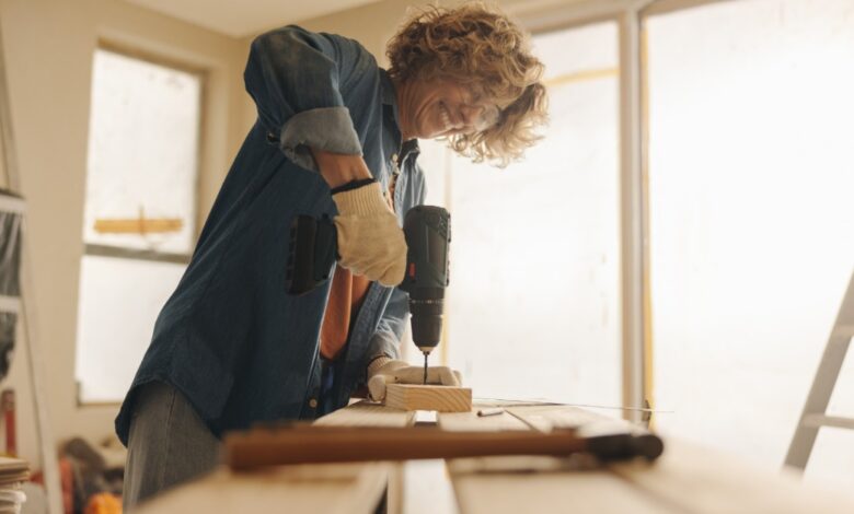 Worker wearing gloves uses a cordless drill on a piece of wood at a workbench inside a bright room under renovation.