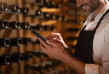 A close-up of the hands of a man holding a smartphone inside a cellar with multiple wine bottles placed on the wall.