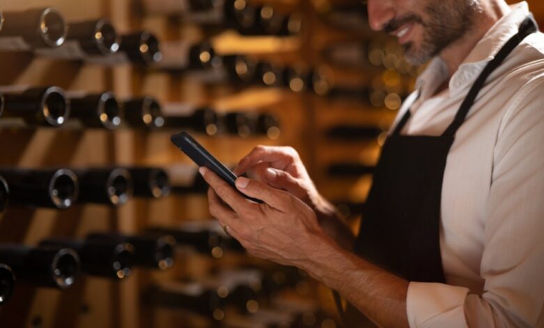A close-up of the hands of a man holding a smartphone inside a cellar with multiple wine bottles placed on the wall.