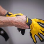 A person is putting on a pair of yellow and black work gloves. The gloves have a strip of Velcro on the top.