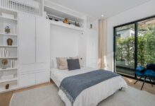 A bedroom with a Murphy bed in front of a large white wall shelf with a closet and multiple décor items on the left.
