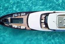 An aerial view of a large, white superyacht floating in turquoise water, with a small lounging pool in the front.