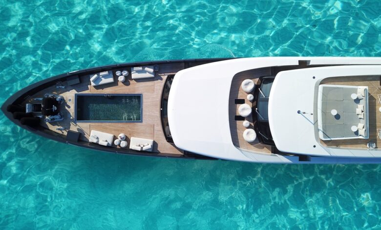 An aerial view of a large, white superyacht floating in turquoise water, with a small lounging pool in the front.