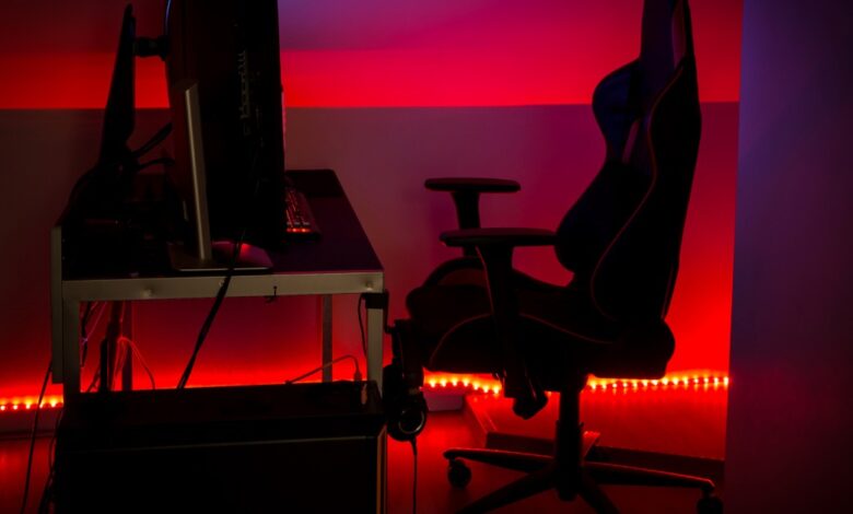 Dark gaming station features a large monitor, ergonomic chair, desk, and vivid red LED lighting along the walls.