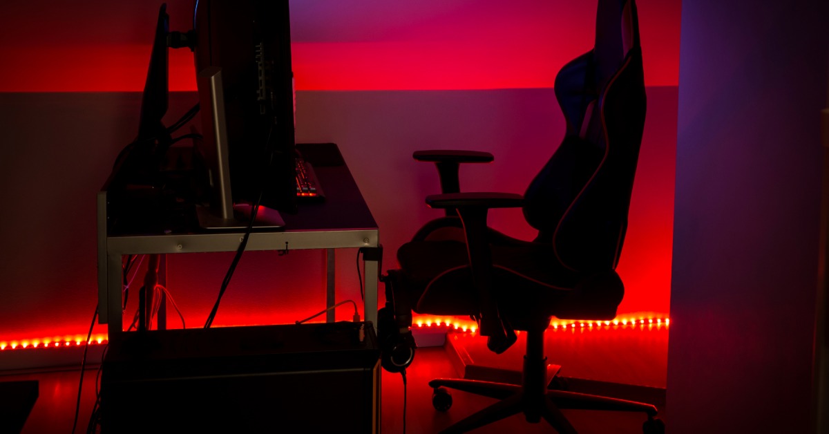 Dark gaming station features a large monitor, ergonomic chair, desk, and vivid red LED lighting along the walls.