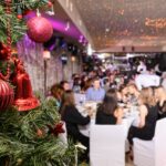 A Christmas tree decorated with red ornaments is in the room with people dressed nicely and sitting at tables.