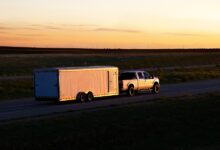 A truck is hauling a trailer down a road. The sun is low on the horizon, causing lowlight conditions.